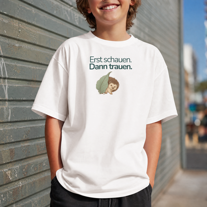 Ein Junge steht vor einer Wand und trägt ein weißes Kinder T-Shirt mit einem Igel und der Aufschrift Erst schauen, Dann trauen.