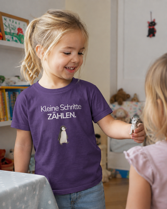 Ein Mädchen steht im Kinderzimmer. Sie trägt ein lila Kinder T-Shirt mit einem Pinguin und dem Spruch Kleine SChritte zählen.