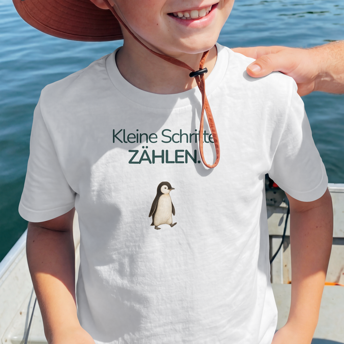 Ein Junge steht in einem Boot und trägt ein weißes Kinder T-Shirt mit einem Pinguin und dem Spruch Kleine Schritte zählen.