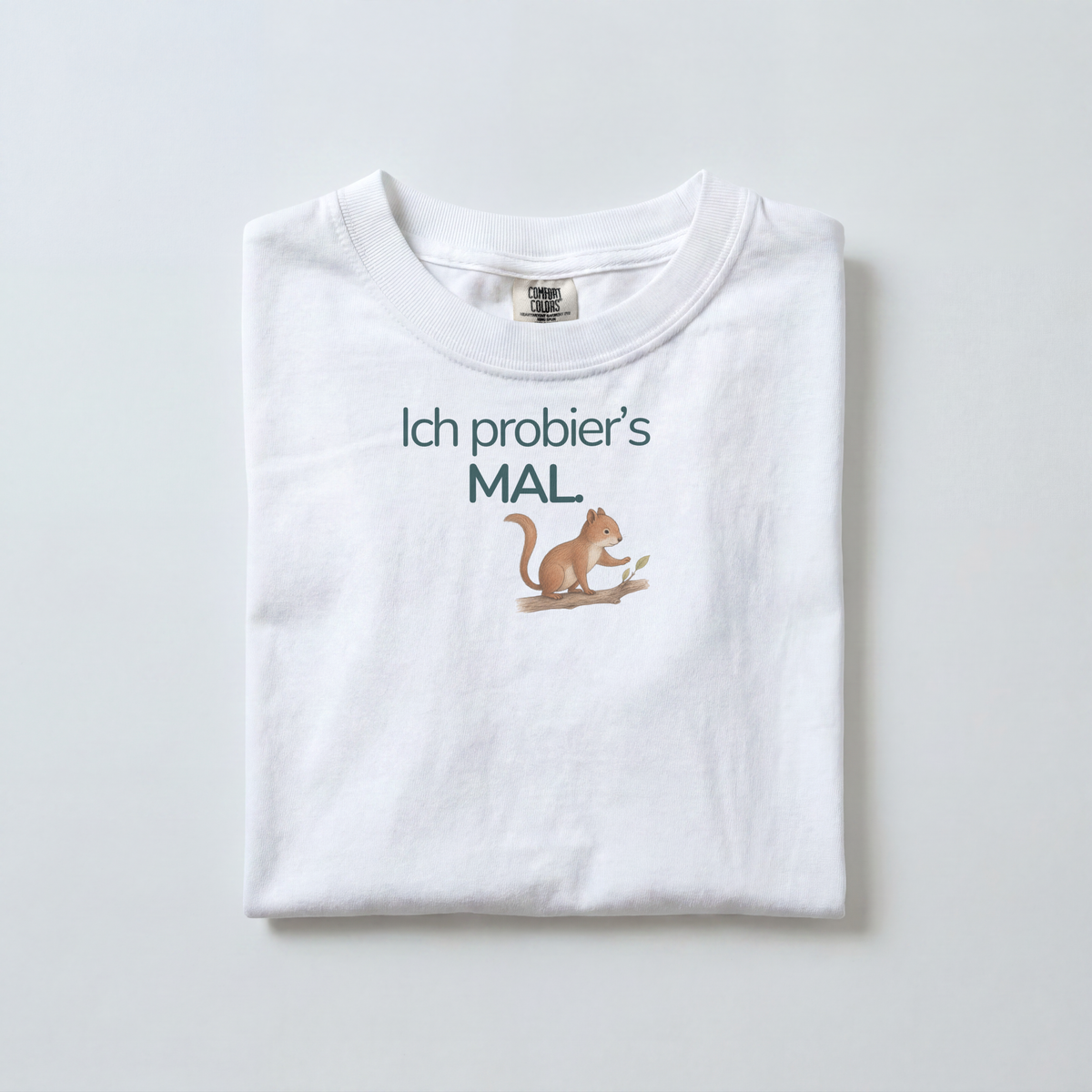 Ein weißes gefaltetes Kinder T-Shirt liegt auf dem Boden. Es zeigt ein Eichhörnchen mit dem Spruch Ich probier's mal