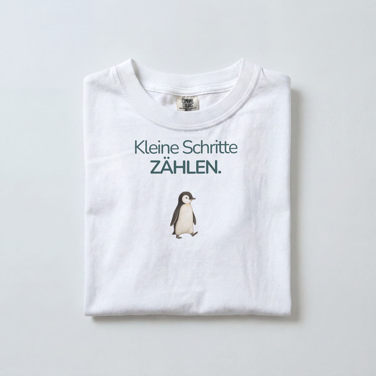 Ein weißes gefaltetes Kinder T-Shirt liegt auf dem Boden. Es zeigt einen Pinguin mit dem Spruch Kleine Schritte zählen