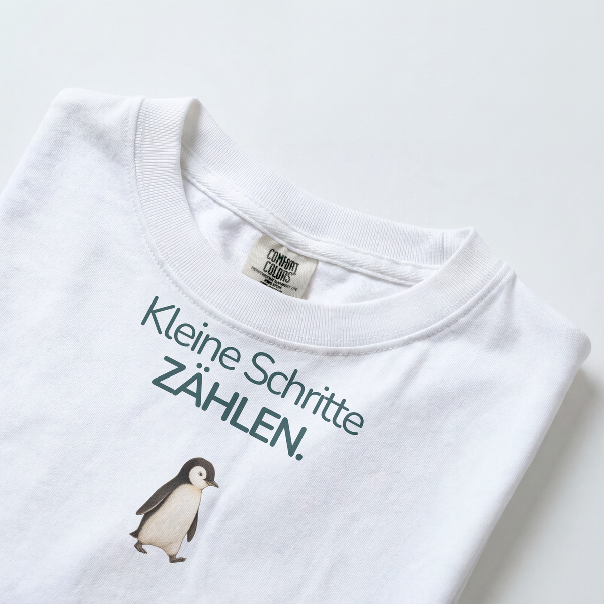 Gezeigt wird der Halsauschnitt eines weißes Kinder T-Shirts mit einem Pinguin und dem Spruch Kleine Schritte zählen