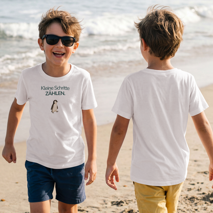 Zwei Jungen sind am Strand. Der linke Junge trägt eine Sonnenbrille und ein Kinder T-Shirt mit dem Spruch Kleine Schritte zählen. Den rechten Jungen sieht man von hinten mit einem Kinder T-Shirt und die Rückseite ist nicht bedruckt