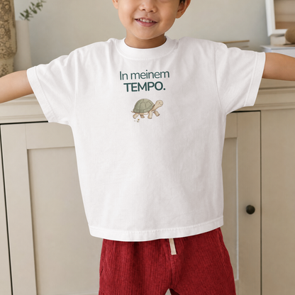 Ein Junge steht wor einem Schrank und trägt ein weißes Kinder T-Shirt mit einer Schildkröte und der Aufschrift In meinem Tempo.