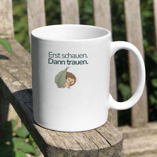 Tasse steht auf einer Armlehne eines Stuhl im Garten. Die asse zeigt einen Igel und sie hat die Aufschrift Erst schauen. Dann trauen.